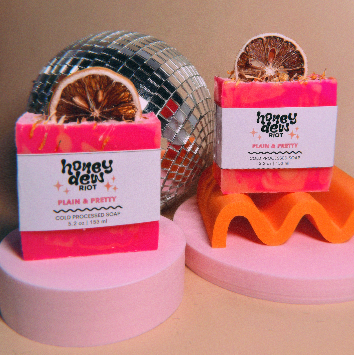 Two pink and orange soap bars decorated with dried orange slices, displayed on pink and orange wavy stands with a disco ball in the background.